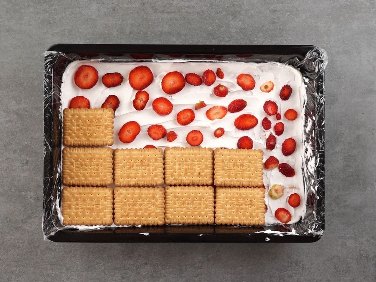 Black baking dish on gray table filled with whipped cream, layers of strawberries, and a layer of crackers