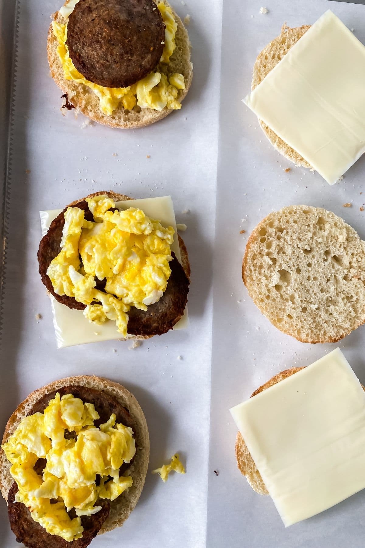 English muffins on baking sheet with sausage and egg on some and cheese on others