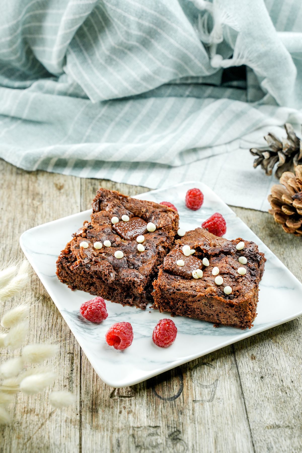 square marble plate holding two brownie slices topped with mini white chocolate chips beside raspberries on wood table with blue striped napkin in background