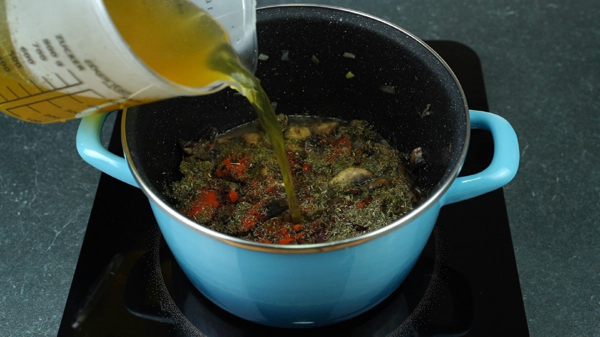 measuring cup of stock being poured into stockpot with mushrooms